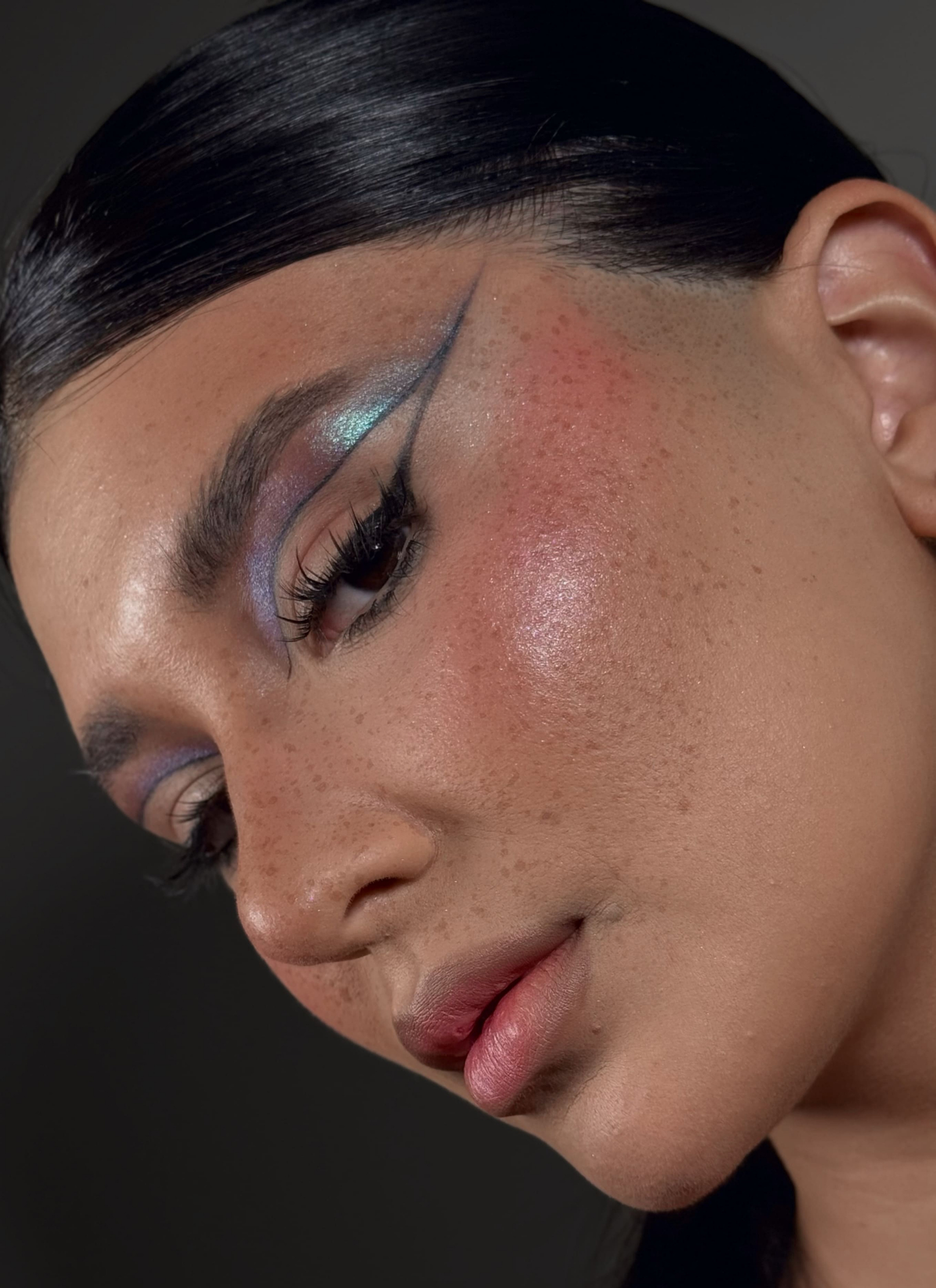 Primer plano de una mujer con maquillaje vibrante en ojos y mejillas, piel con pecas y cabello oscuro recogido.