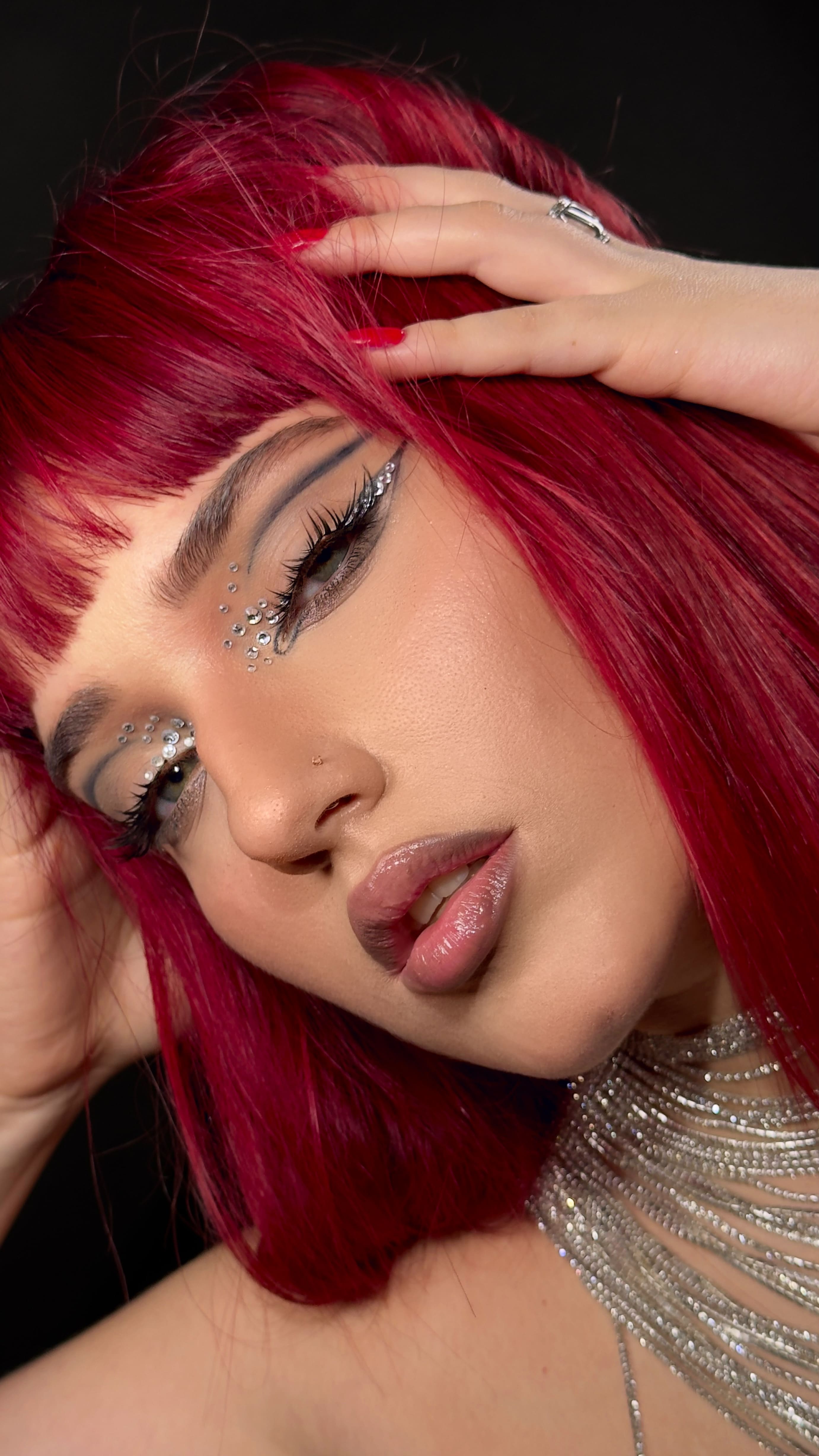 Primer plano de una persona con cabello largo y rojo brillante, maquillaje con piedras brillantes alrededor de los ojos y collar plateado brillante.