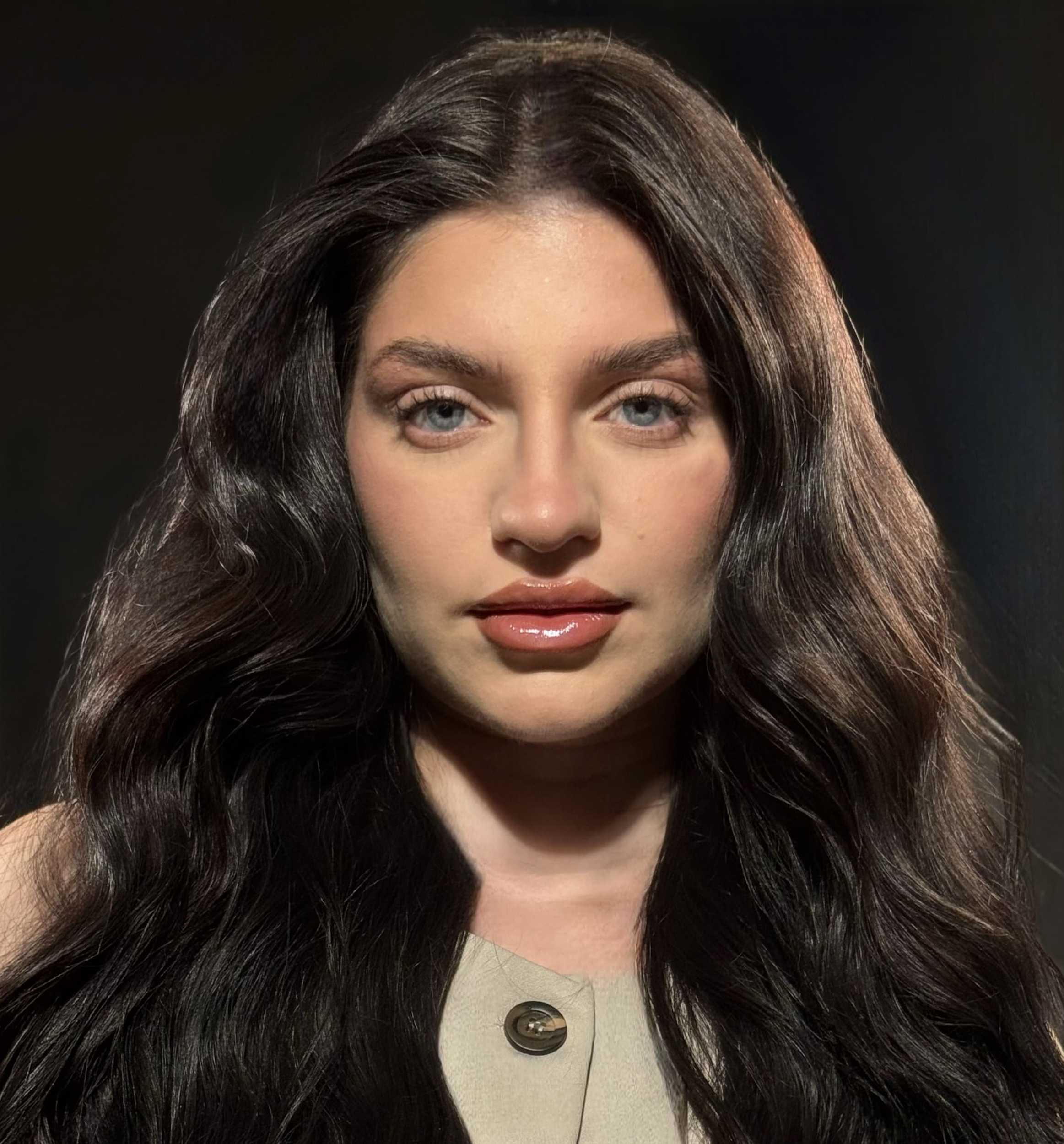 Primer plano de una mujer con cabello largo y ondulado, ojos claros y labios brillantes, sobre fondo oscuro.