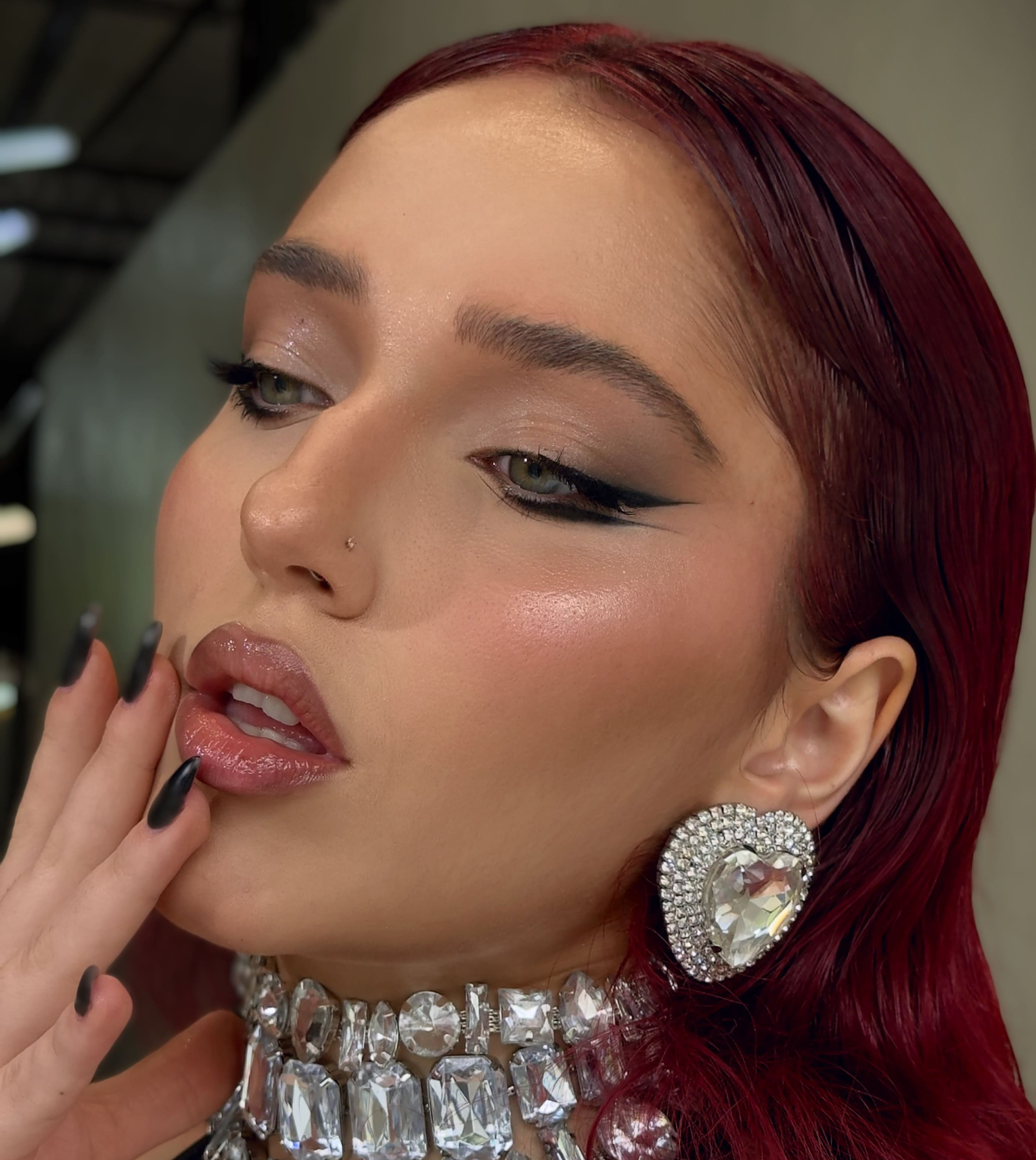 Primer plano de mujer con maquillaje de ojos ahumados, labios brillantes, arete en forma de corazón con pedrería y collar brillante.